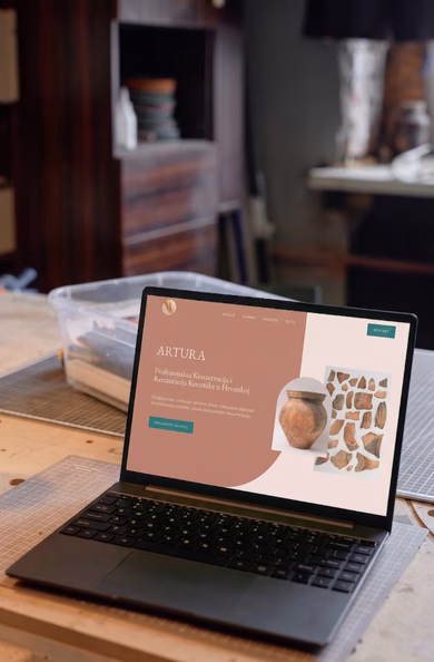 Ein geöffneter Laptop steht auf einer hölzernen Werkbank in einem Atelier und zeigt die neu gestaltete, erdfarbene Website von Artura zum Thema Keramik-Restaurierung in Kroatien.