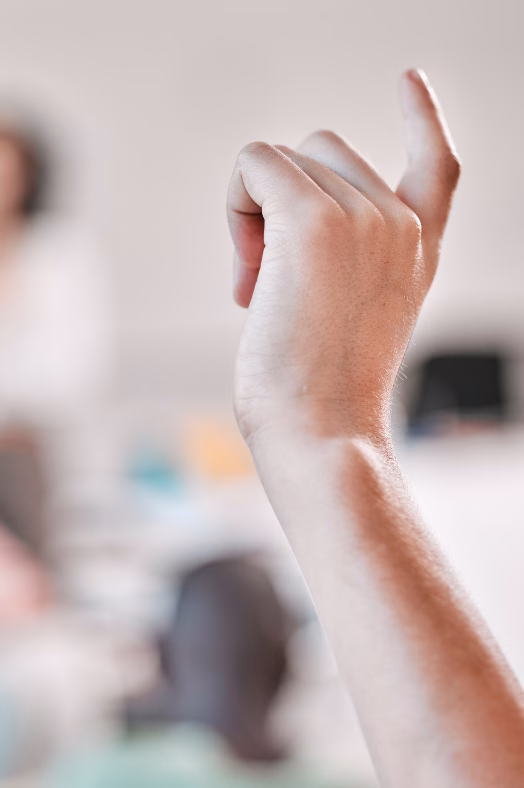 Eine Hand im Vordergrund ist mit erhobenem Zeigefinger leicht nach oben gestreckt, im unscharfen Hintergrund ist eine Beratungssituation im Büro zu erkennen.