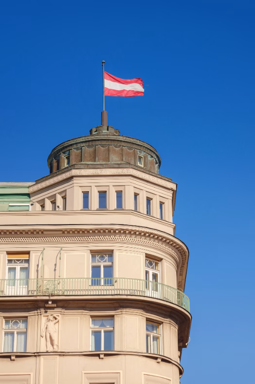 Die Kuppel eines klassischen Wiener Altbaus mit einer wehenden rot-weiß-roten Österreich-Fahne vor klarem blauen Himmel.