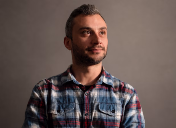 Porträt von Stefan, Experte für Online-Marketing-Strategie und Suchmaschinenoptimierung, im karierten Hemd vor grauem Hintergrund.